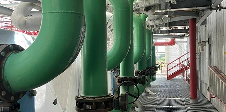 Industrial facility with large green-painted pipes running through the structure, with valves and steel grating flooring visible.