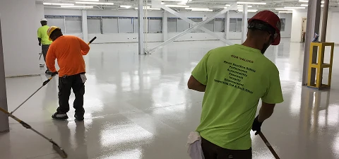 Construction crew applying epoxy flooring with rollers across a large industrial space, creating a smooth glossy surface.