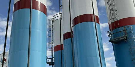 Row of tall industrial silos painted in white and blue with red accent bands, equipped with ladders and access platforms.