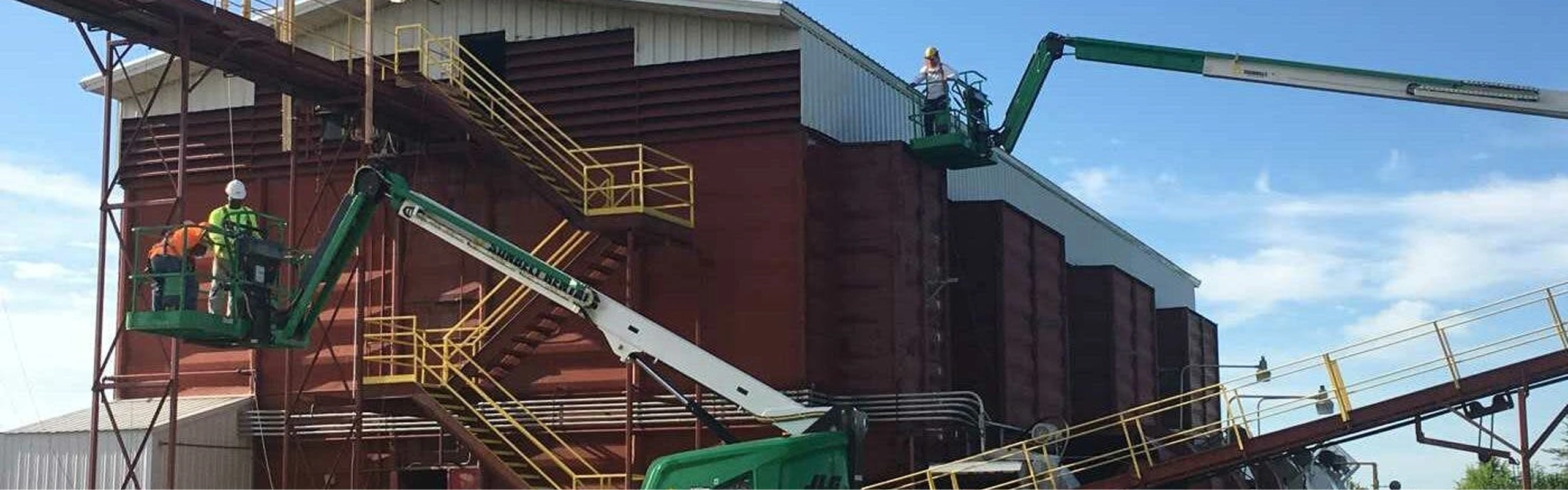 Three construction workers wearing safety gear operate two green boom lifts to access the exterior of a large red industrial building with yellow staircases and metal structures under a blue sky.