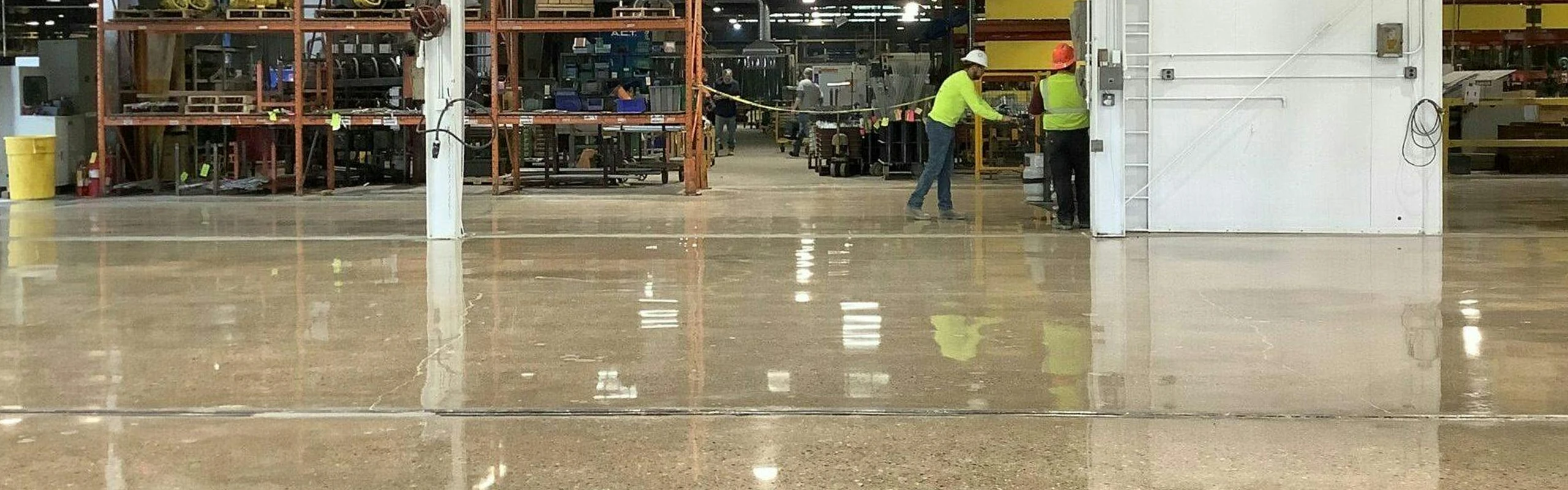 Polished concrete production area with workers in safety gear near a white wall; shelving and machinery in the background.