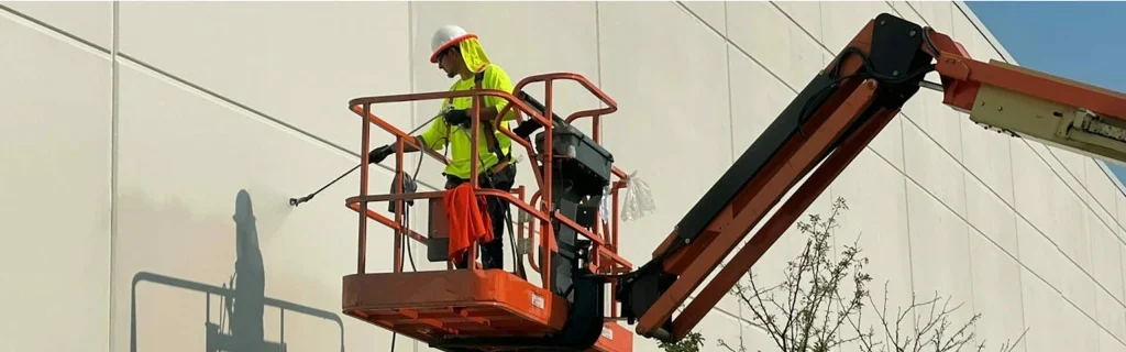 Worker in safety gear on a boom lift spraying and painting the exterior wall of a large commercial building.
