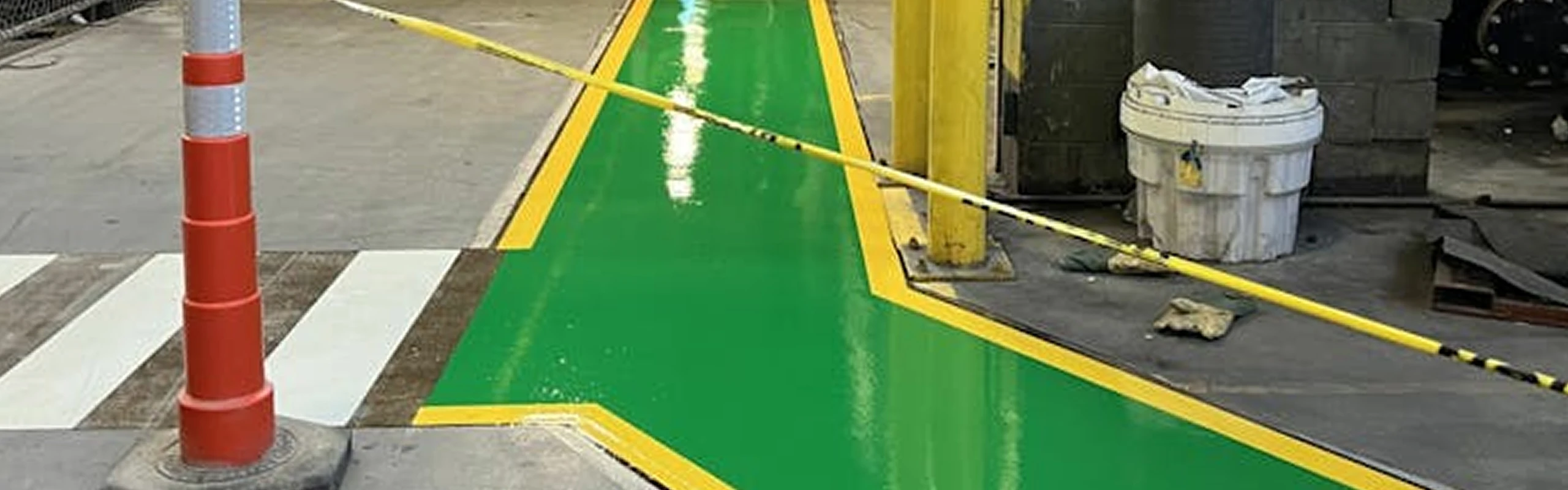 Fresh green pedestrian walkway with yellow borders painted on a factory floor at Pilkington, cordoned with caution tape; cone, zebra crosswalk, and yellow columns nearby.