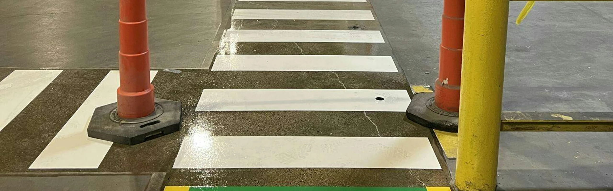 Fresh white zebra-style crosswalk stripes on factory concrete between orange cones and a yellow guardrail.