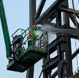 Worker using a lift to power wash a large steel structure outdoors, spraying water at high pressure.