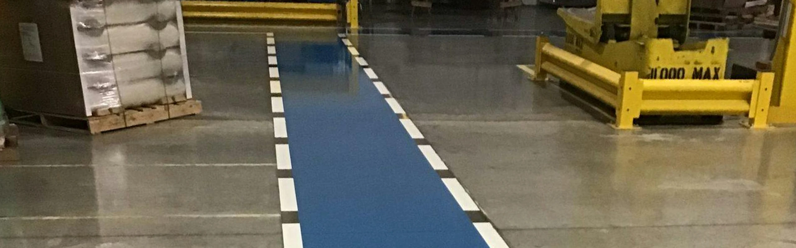 Blue pedestrian walkway with dashed white borders painted on a polished warehouse floor at Wrisco Industries Inc, with yellow safety rails and pallets nearby.