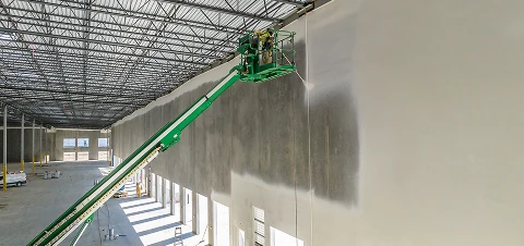 Worker on a green boom lift spraying or painting a very tall interior warehouse wall beneath an open metal roof.