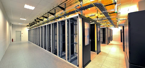 Wide aisle in a modern data center lined with tall server racks and overhead cable trays in a brightly lit room.