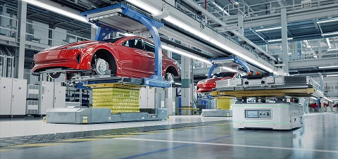 Red electric vehicles on an automated factory line, each raised on yellow platforms with robotic equipment and overhead lighting.